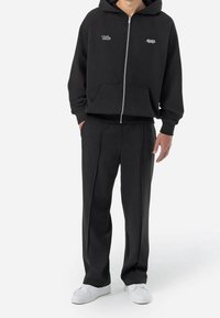Felpa nera con cerniera e tasche frontali, con polsini a costine, abbinata a pantaloni neri dritti, indossata con sneakers bianche.