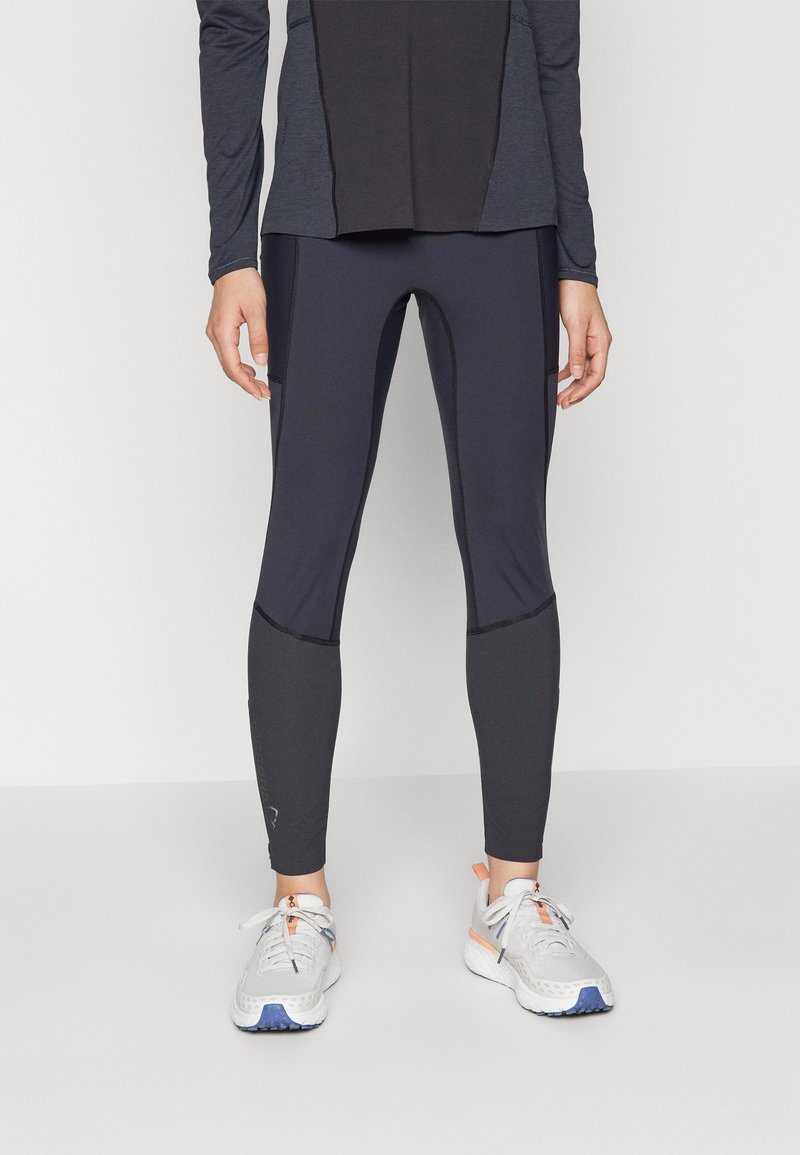 Leggings atletici neri con accenti grigi a contrasto, vita media, tessuto elasticizzato, abbinati a sneakers grigie chiare con dettagli arancioni.