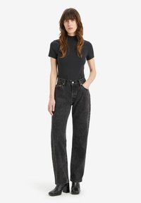 Zwarte hoge taille jeans met een vervaagde textuur, gecombineerd met een strak zwart t-shirt. Het model heeft lang golvend haar en draagt chunky zwarte hakken.
