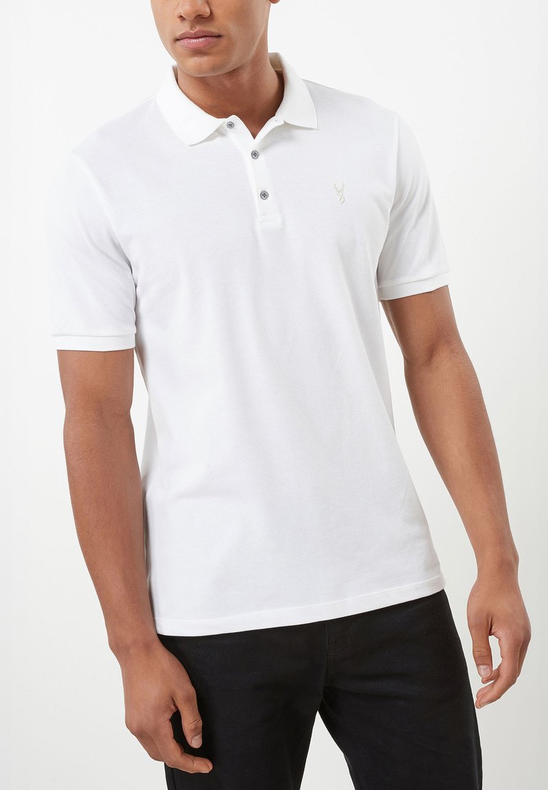 Homme portant un polo blanc à manches courtes avec trois boutons et un petit logo sur la poitrine, associé à un pantalon noir.