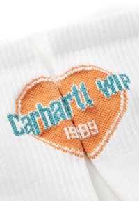 Chaussettes blanches avec un grand design de cœur orange. Le texte en turquoise indique "Carhartt WIP" et "1989" dans une police audacieuse et claire.