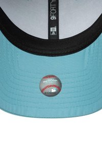 Primer plano de la visera de una gorra azul claro y negra con una etiqueta holográfica oficial de las Grandes Ligas de Béisbol y un número de serie.