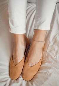 Bailarinas de ante color beige con un diseño bajo en forma de V. Textura suave y punta redondeada. Llevadas con pantalones recortados blancos sobre un fondo claro.