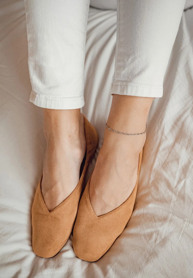 Bailarinas de ante color beige con un diseño bajo en forma de V. Textura suave y punta redondeada. Llevadas con pantalones recortados blancos sobre un fondo claro.