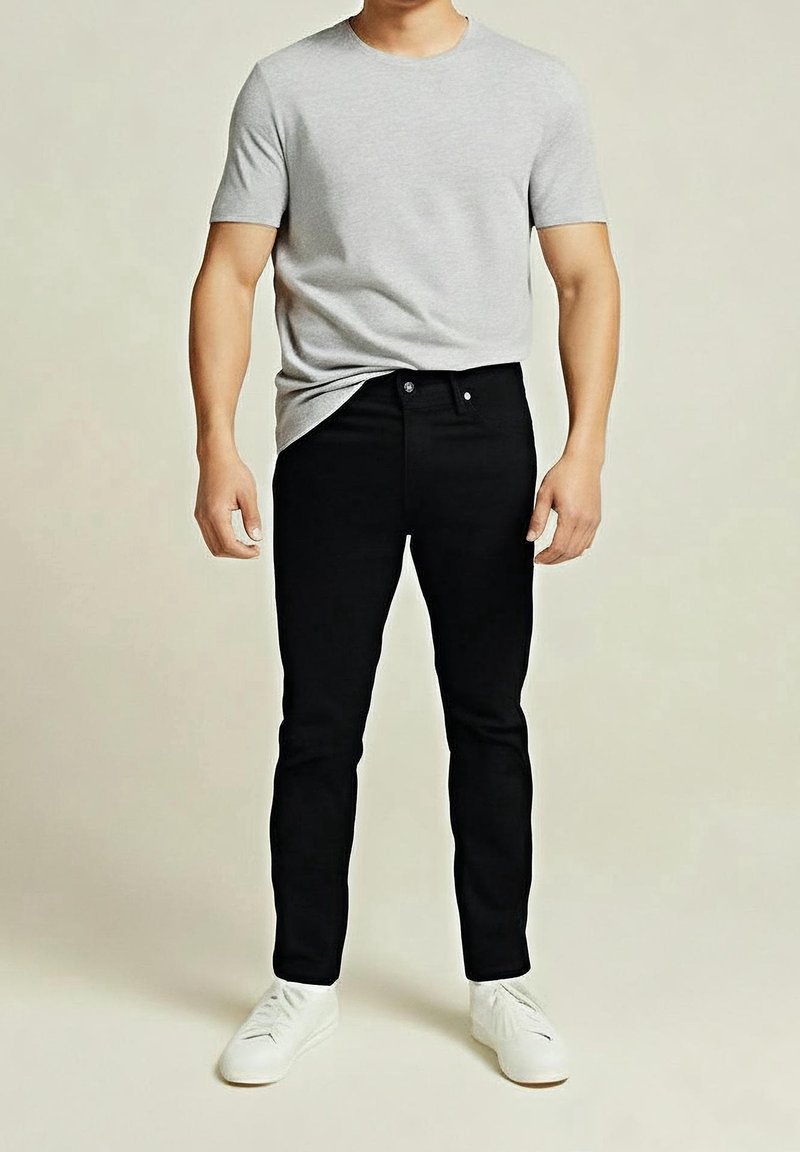 Homme portant un t-shirt gris clair à manches courtes, un jean slim noir et des baskets blanches, debout devant un fond uni.