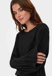 Sort plisseret top med tekstureret stof, lange ærmer og rund halsudskæring, der har markante plisseringer og en afslappet pasform.