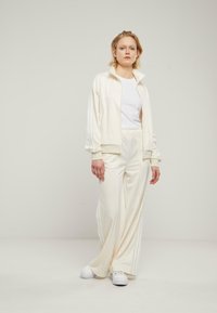 Creme tracksuit sett laget av glatt stoff, med en glidelåsjakke med ståkrage og vide bukser, begge med hvite side-striper.