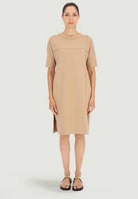 Beżowa sukienka T-shirt z krótkimi rękawami, okrągłym dekoltem, bocznymi rozporkami i gładką teksturą. Noszona z czarnymi sandałami z złotymi akcentami.