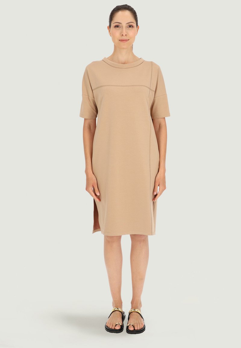 Beżowa sukienka T-shirt z krótkimi rękawami, okrągłym dekoltem, bocznymi rozporkami i gładką teksturą. Noszona z czarnymi sandałami z złotymi akcentami.