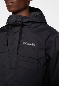 Černá bunda Columbia s kapucí, se dvěma předními kapsami, texturovanou látkou a nastavovacími šňůrkami u límce.