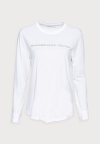 Witte lange mouwen shirt gemaakt van zachte stof, met een ronde halslijn en een lichtgrijs logo op de borst. De zoom is iets gebogen.