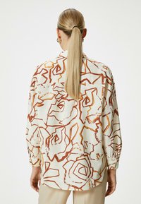 Long-sleeve, loose-fitting shirt made of light fabric, featuring a cream background with abstract brown floral patterns and gold accents.