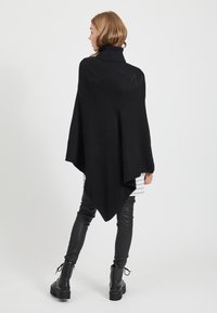 Poncho de malha preto com colarinho alto e barra assimétrica. Apresenta uma textura suave e um design solto e drapeado. Usado sobre uma blusa riscada e calças de couro.