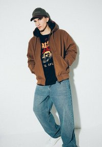 Brązowa kurtka z kapturem na czarnej koszulce z grafika, w połączeniu z luźnymi niebieskimi dżinsami i białymi sneakerami. Model nosi zieloną czapkę baseballową.
