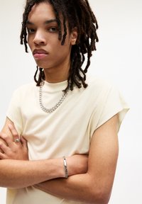 Lichtbeige, kortemouwen t-shirt met een relaxed fit en ronde halslijn. Zilveren ketting met een armband om de arm, structuurrijk haar.