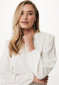 Camicia bianca con motivi floreali ricamati, maniche lunghe a sbuffo e polsi elasticizzati. Il modello ha i capelli lunghi e uno sfondo neutro.