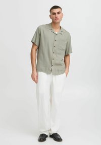 Camisa de botón de manga corta en color oliva con un bolsillo, combinada con pantalones de lino blancos sueltos y zapatos negros. Tela suave, corte relajado.