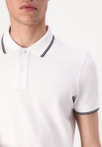 Biela polo košeľa z jemnej tkaniny s dvojitým gombíkovým zapínaním, vyšitým logom a golierom a manžetami v námorníckych pruhovaných farbách.