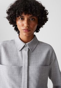 Camicia grigia con bottoni realizzata in tessuto testurizzato, presenta un colletto a punta e due tasche frontali, progettata con una vestibilità ampia e dettagli minimi.