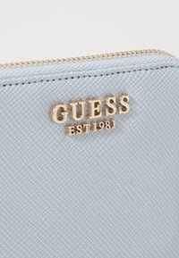 Porte-monnaie zippé texturé bleu clair avec un logo GUESS en doré et l'inscription "EST 1981". Motif de surface détaillé visible.