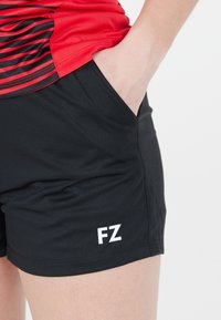 Person i svarta sportshorts med "FZ"-logotyp och en röd och svart randig sporttröja, med handen placerad i shortsfickan.
