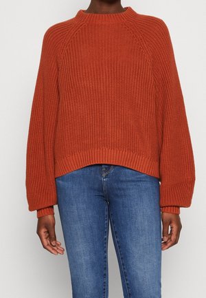 Osoba nosící rezavě oranžový silný pletený svetr s balónovými rukávy v kombinaci se středně modrými džínami na jednobarevném pozadí.