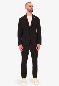 Blazer nero su misura con rever a punta, doppie tasche frontali, abbinato a pantaloni slim-fit coordinati e sneakers beige. Texture del tessuto liscia.