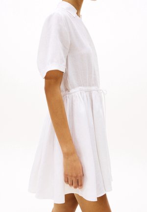 Robe blanche courte avec un col et des manches courtes. Elle est dotée d'une taille ajustable par cordon et d'une jupe fluide en tissu de lin.