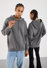 Felpa grigia con cappuccio, tasca frontale, chiusura a coulisse e polsini a coste. Il modello indossa jeans azzurri; un altro modello in nero sotto.