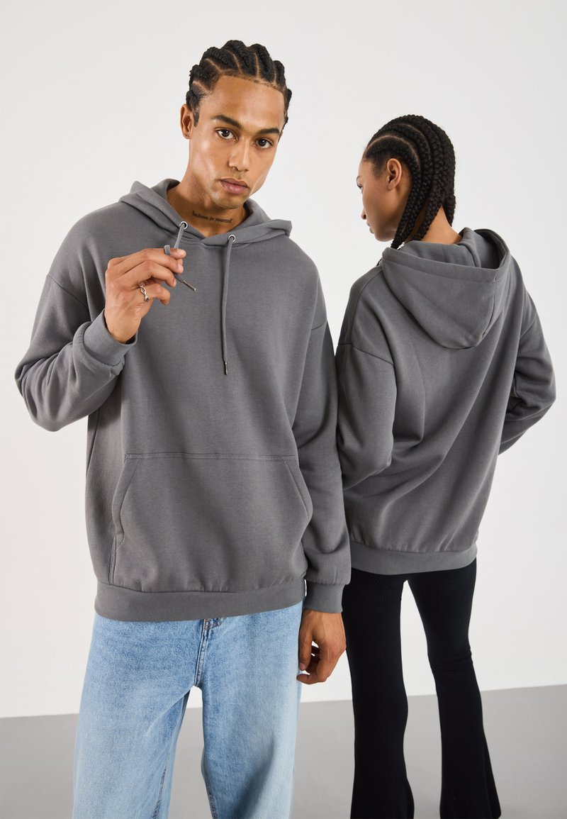 Felpa grigia con cappuccio, tasca frontale, chiusura a coulisse e polsini a coste. Il modello indossa jeans azzurri; un altro modello in nero sotto.