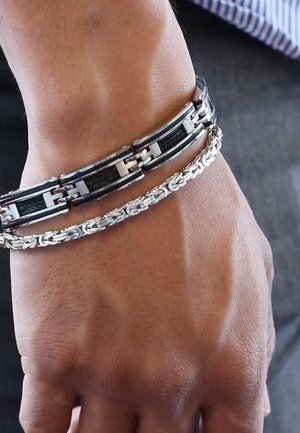 Deux bracelets sur un poignet : une chaîne en argent avec un motif tissé et un bracelet en métal noir avec des accents argentés brillants, le tout sur un fond de peau texturée.