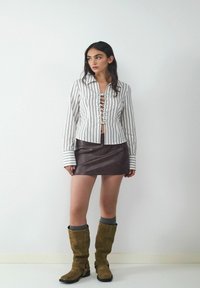Gestreept wit shirt met vetersluiting aan de voorkant, gecombineerd met een bruin leren minirok en olijfgroene kniehoge laarzen. Grijze sokken zichtbaar.