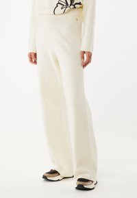 Pantalones blancos de pierna ancha, hechos de una tela suave y texturizada; cuentan con una cinturilla elástica y un pequeño logo de acento dorado.