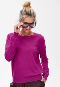 Kobieta z blond włosami w kokiem nosi fioletowy sweter i brązowe spodnie, poprawiając duże okulary w musztardowym kolorze, na tle białego tła.