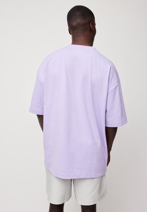 Homme aux cheveux courts portant une chemise oversize manches courtes violet clair et un short de couleur claire, vu de dos sur un fond uni.