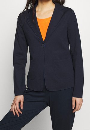 Femme portant un blazer bleu marine ajusté avec deux poches avant sur un haut orange, assorti d'un pantalon bleu marine, debout avec une jambe pliée.