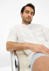 Camicia bianca di lino a maniche corte con colletto alla cinese, dotata di una tasca sul petto. Modello seduto su una poltrona in cromato.