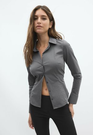 Jeune femme aux longs cheveux bruns porte une chemise grise à fines rayures partiellement déboutonnée et un pantalon noir devant un fond clair uni.