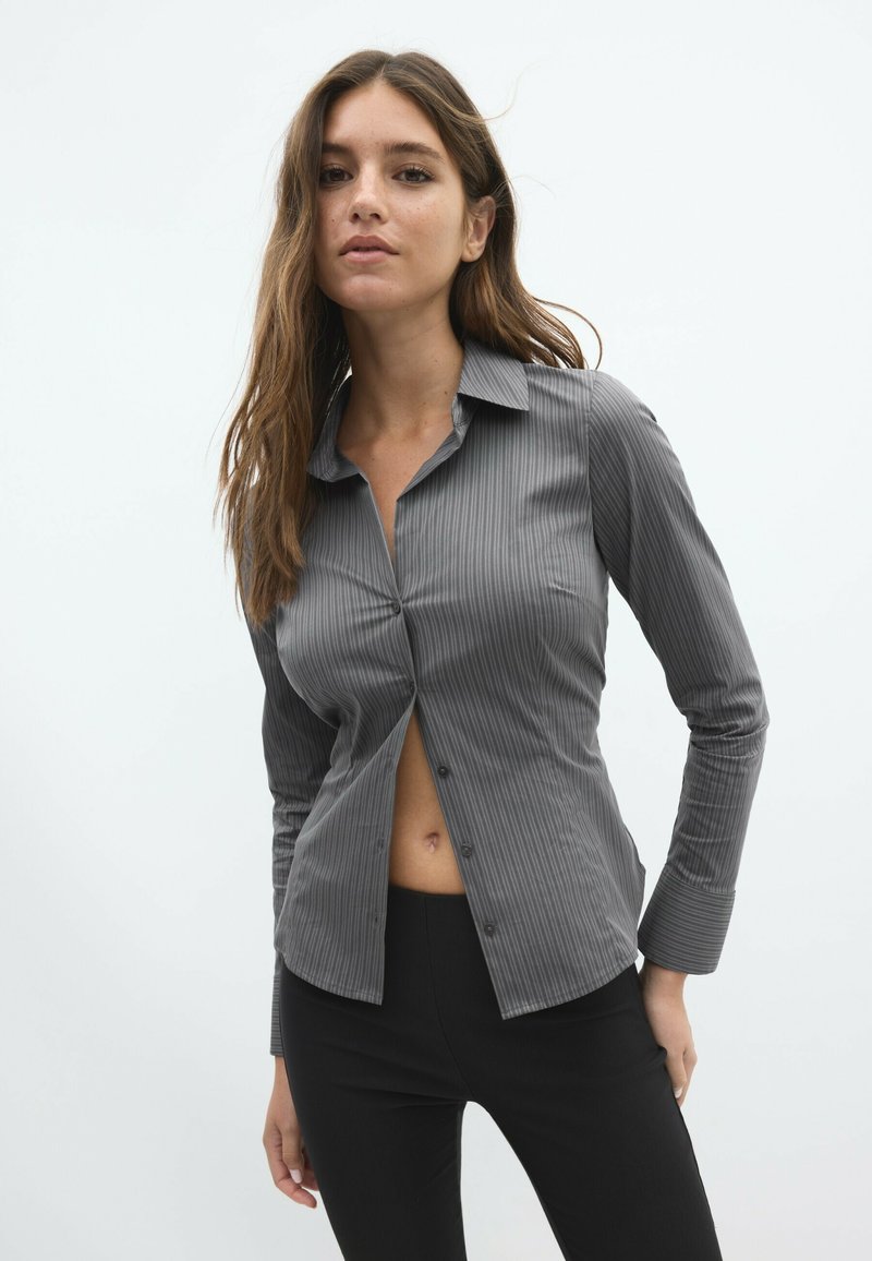 Jeune femme aux longs cheveux bruns porte une chemise grise à fines rayures partiellement déboutonnée et un pantalon noir devant un fond clair uni.
