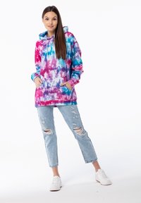 Il felpa con cappuccio presenta un motivo tie-dye multicolore in rosa, blu e viola, con una tasca frontale. Abbinata a jeans in denim chiaro a look usurato.