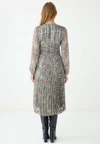 Vrouw in een langlopende, geplooide jurk met luipaardprint, gecombineerd met zwarte kniehoge laarzen, staand tegen een effen witte achtergrond.