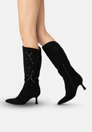 Sorte ruskindsknæhøje støvler med udskårne blomster mønstre og slanke hæle båret på krydsede ben.