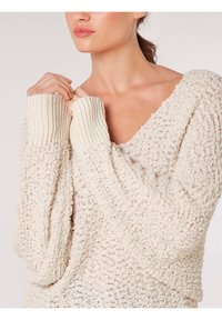 Jasnobeżowy sweter z dzianiny z dekoltem w serek i prążkowanymi mankietami. Posiada teksturowany wzór oraz luźne, zwisające rękawy dla wygody.