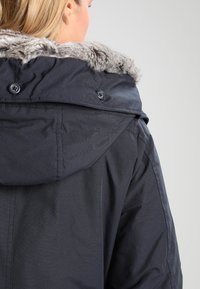 Ciemnoniebieska kurtka z kapturem wyłożonym futrem, zapinana na guziki, wykonana z teksturowanej tkaniny. Widok z tyłu ukazuje kołnierz i projekt ramion.