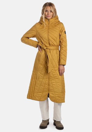 Naine kannab sinepikollast kapuutsiga ja vööga pikemat tikitud mantlit, valgeid pükse ja pruune pahkluu-saapaid, seistes ühe käega puusal.