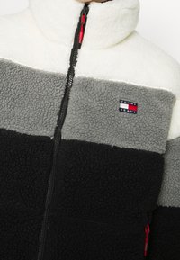 Giacca in fleece con pannelli bianchi, grigi e neri, colletto alto, chiusura con zip, toppa con logo Tommy Jeans e dettagli rossi sulla zip.