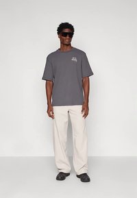 Homem com uma t-shirt oversized cinza escura, calças largas bege claras, sapatos pretos e grandes óculos de sol pretos, de pé contra um fundo simples.