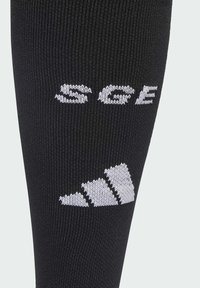 Calzino sportivo nero realizzato in un materiale a maglia testurizzato e elastico. Presenta la scritta bianca "SCE" e un logo con tre strisce vicino alla parte superiore.