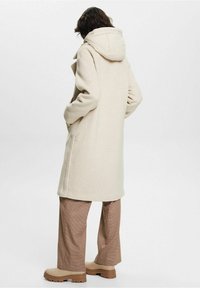 Abrigo de lana color beige con capucha, con un corte recto y bolsillos laterales. Combinado con pantalones marrones de cuadros y botas gruesas beige.