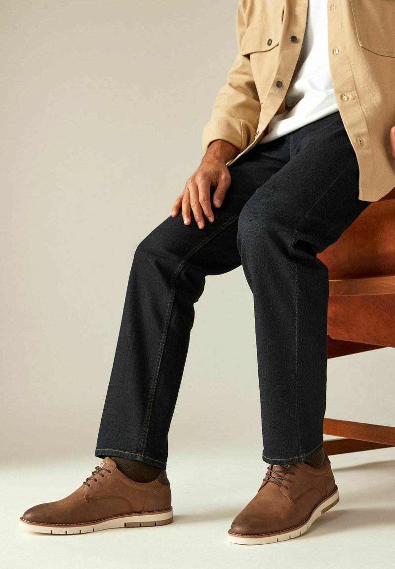 Chaussures en cuir marron avec une semelle blanche, portées avec un jean en denim foncé et une chemise boutonnée beige clair. Détails de design texturés et ajustés visibles.